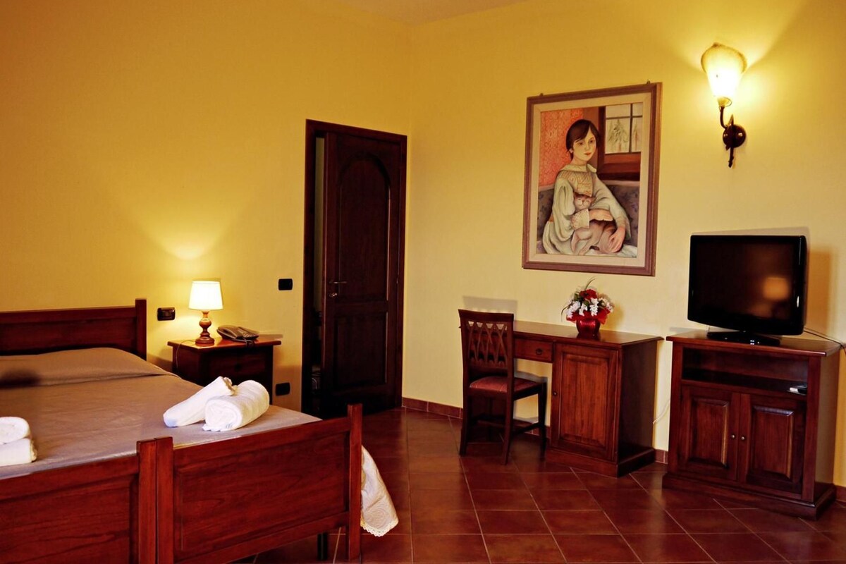 A hotel room with a bed, bedside table with lamp, desk with chair, television, and a painting on the wall.