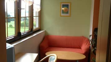 Una habitació amb un sofà vermell, una taula rodona petita, una cadira, un escriptori i una finestra amb cortines.