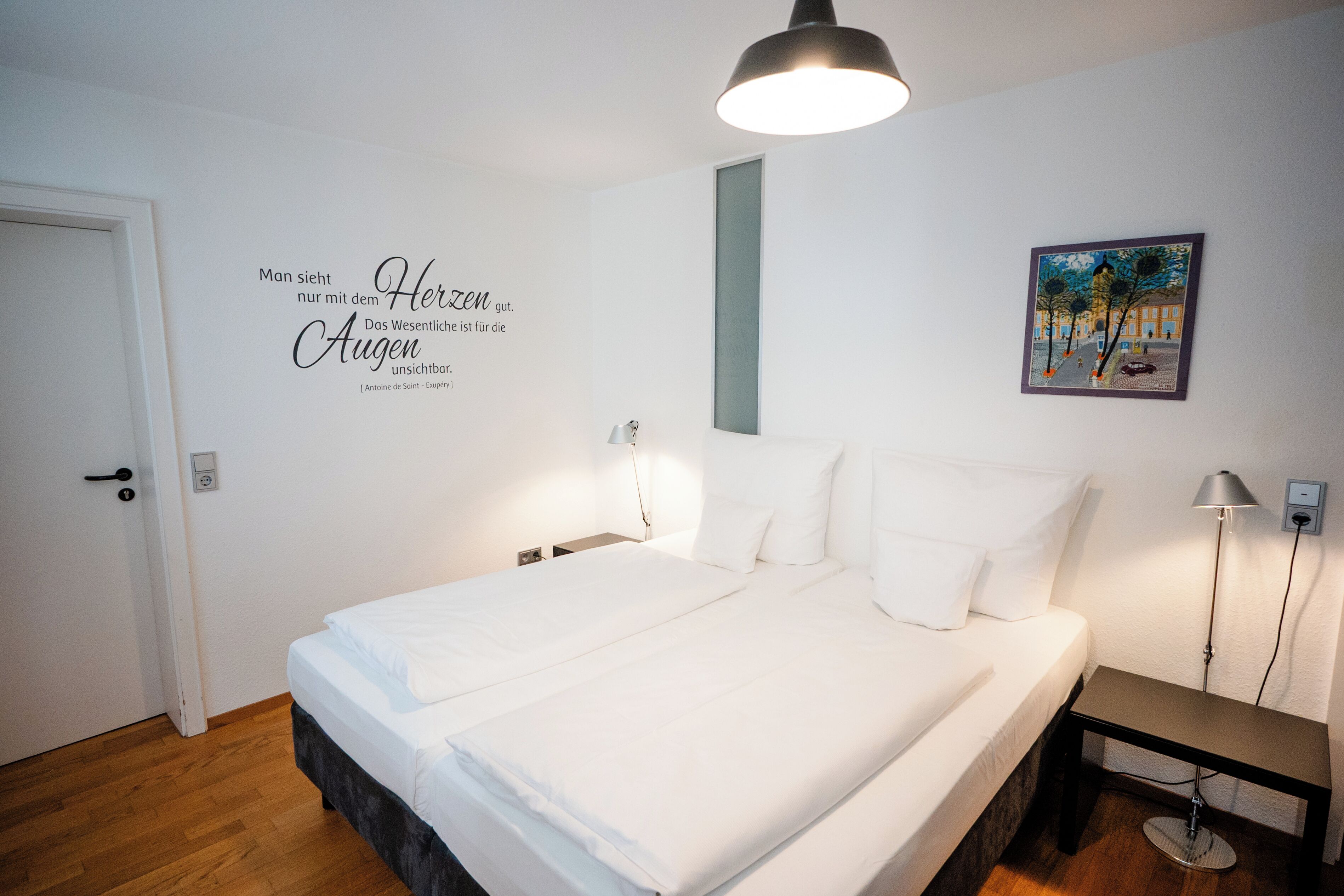 Une chambre à coucher avec un lit, des lampes de chevet, une citation sur le mur, une porte et un tableau.