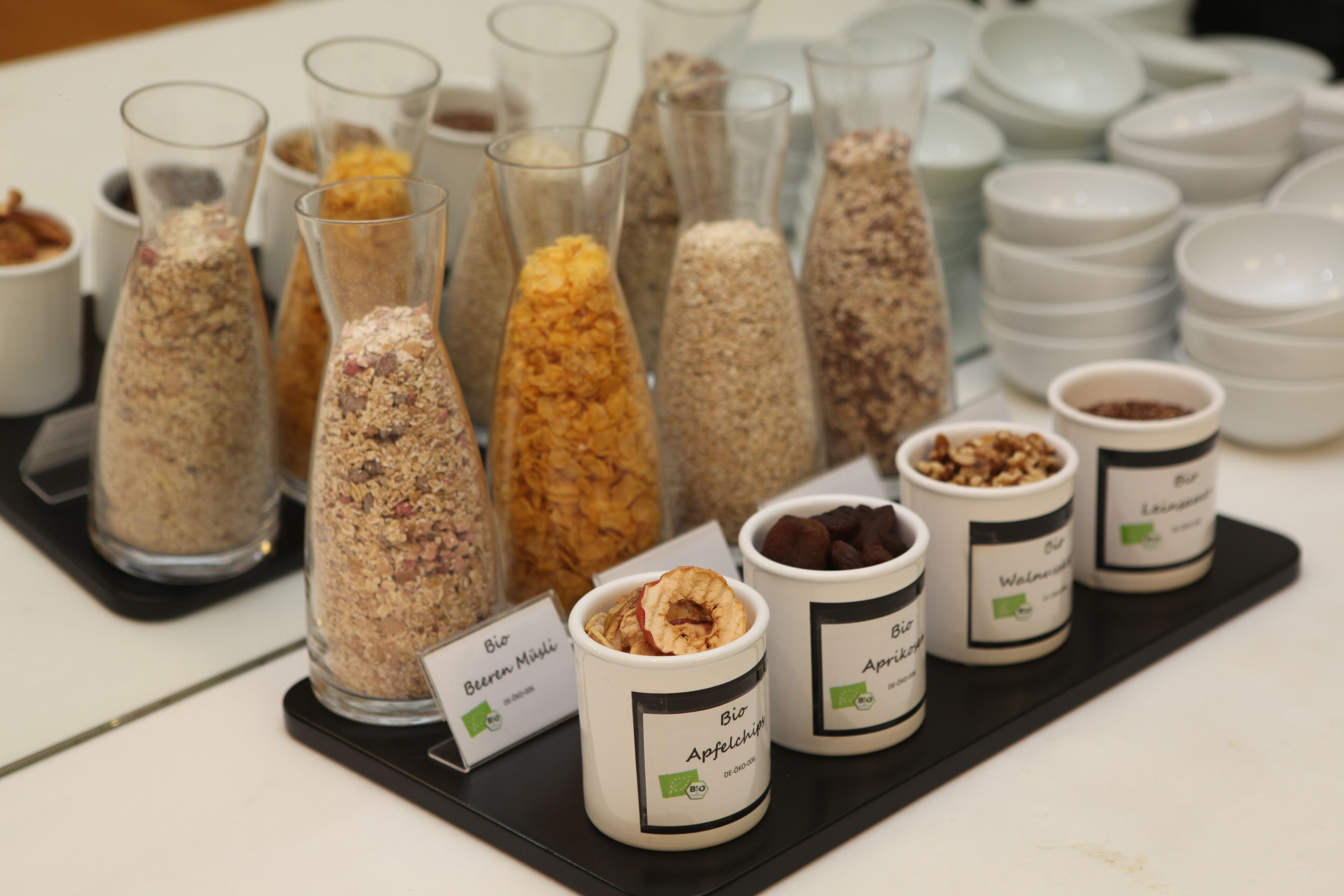 Diversos alimentos expuestos en recipientes de vidrio y tazas de cerámica, cada uno con su nombre e ingredientes.