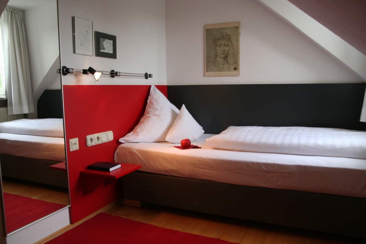 A bedroom with two beds, a red accent wall, a bedside table with a book, and a painting on the wall.