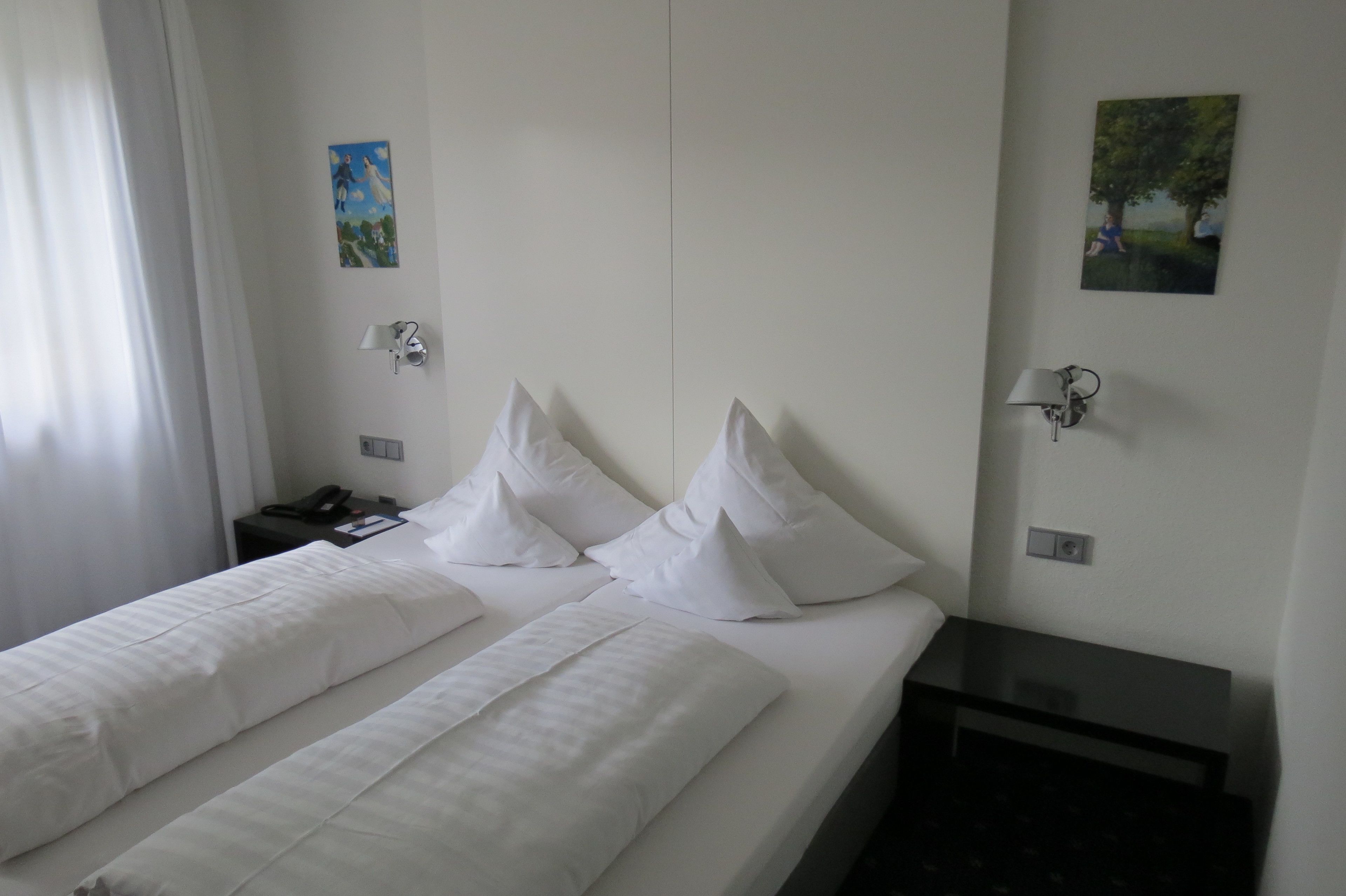 A hotel room with two beds, white bedding, a nightstand with a lamp, and two wall-mounted lamps.