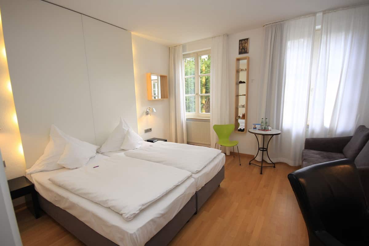 A hotel room with a double bed, a small table, a green chair, a sofa, and a window with curtains.