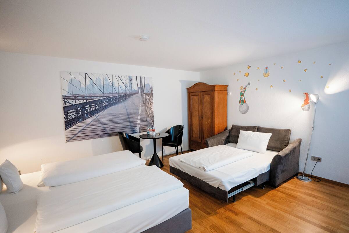 A hotel room with two beds, a wooden cabinet, a small table with chairs, a wall-mounted lamp, and a cityscape painting.