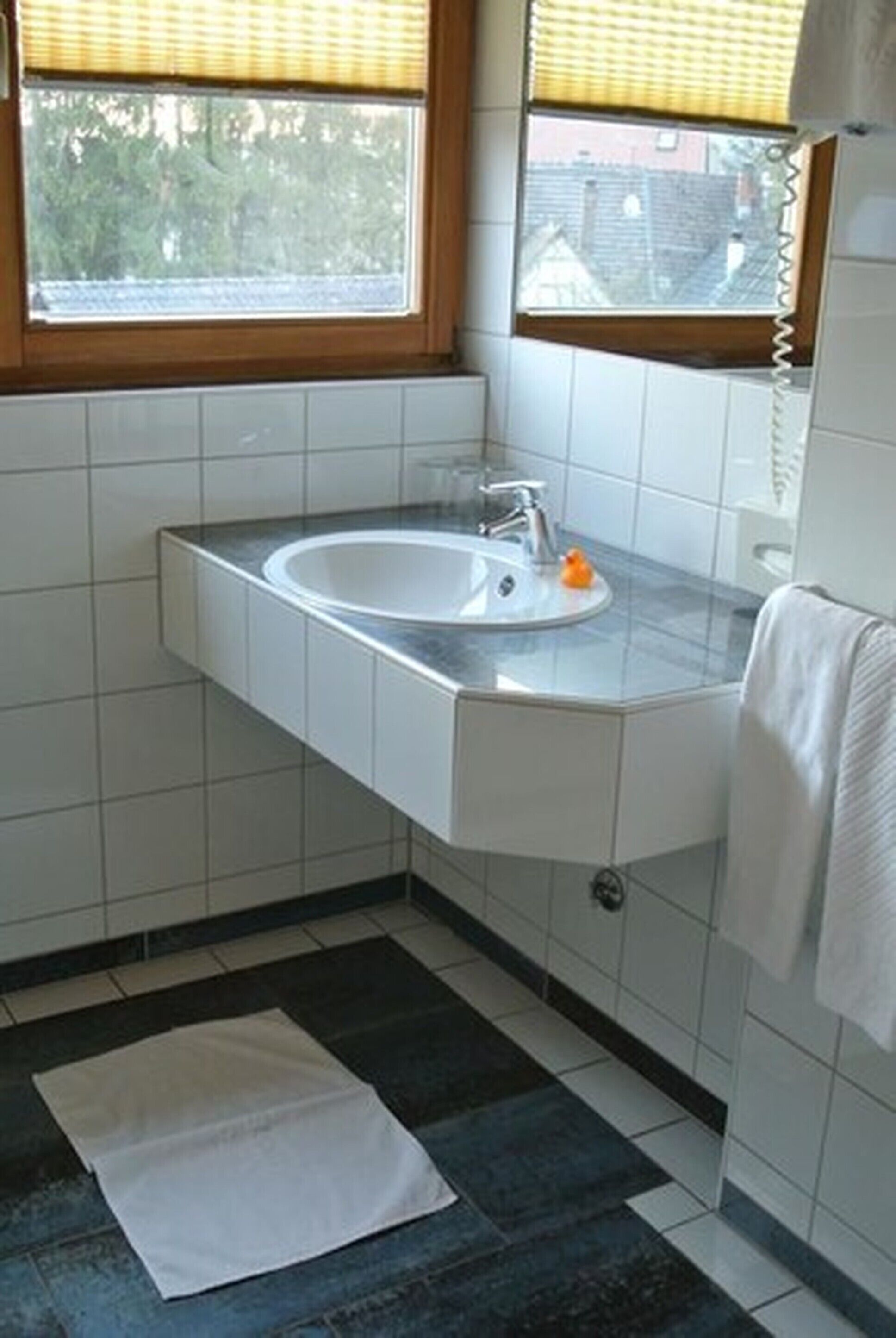 A bathroom with a corner sink, a window with blinds, and a white towel hanging on the wall.