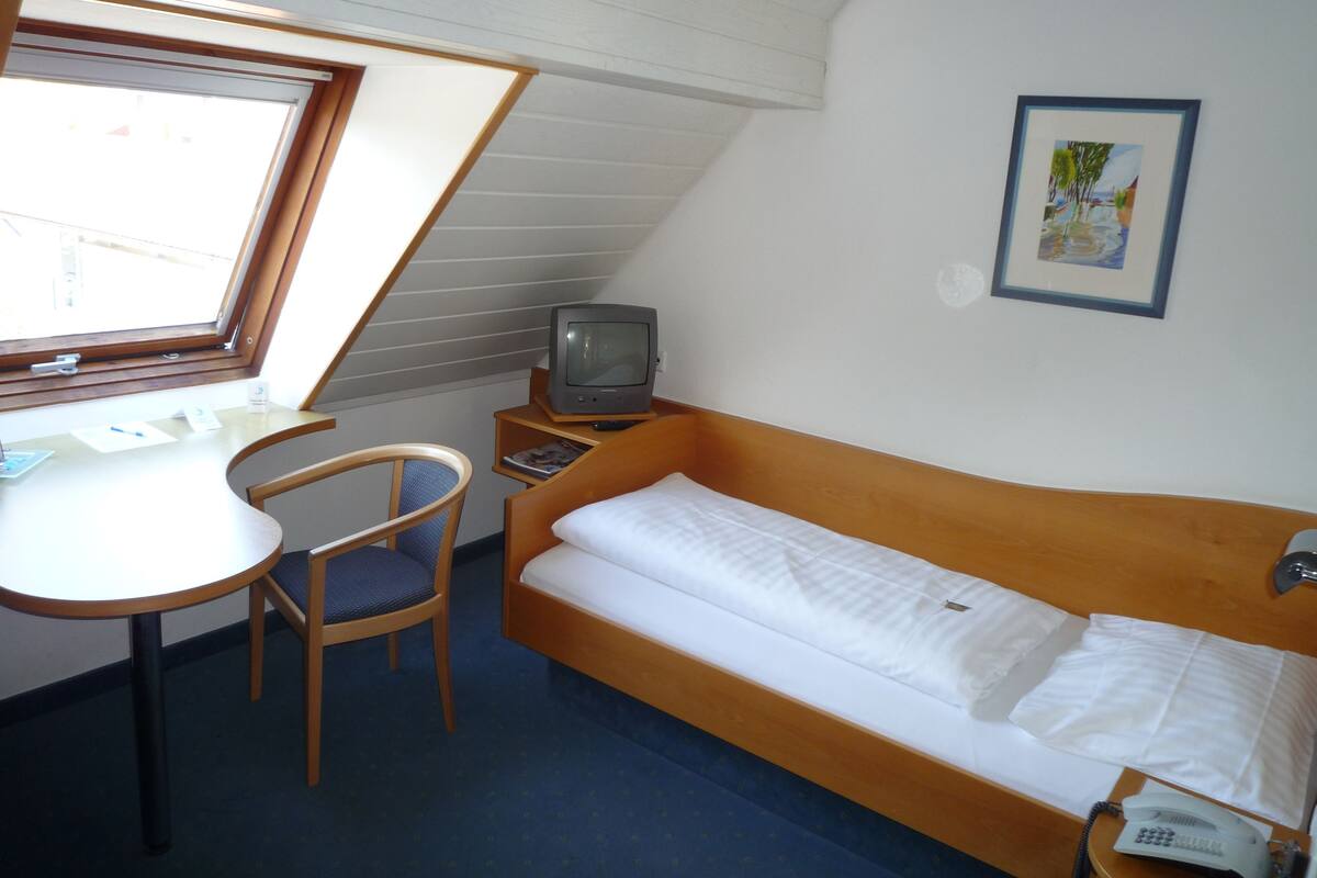 A small, compact room with a bed, a desk, a chair, a television, and a phone.