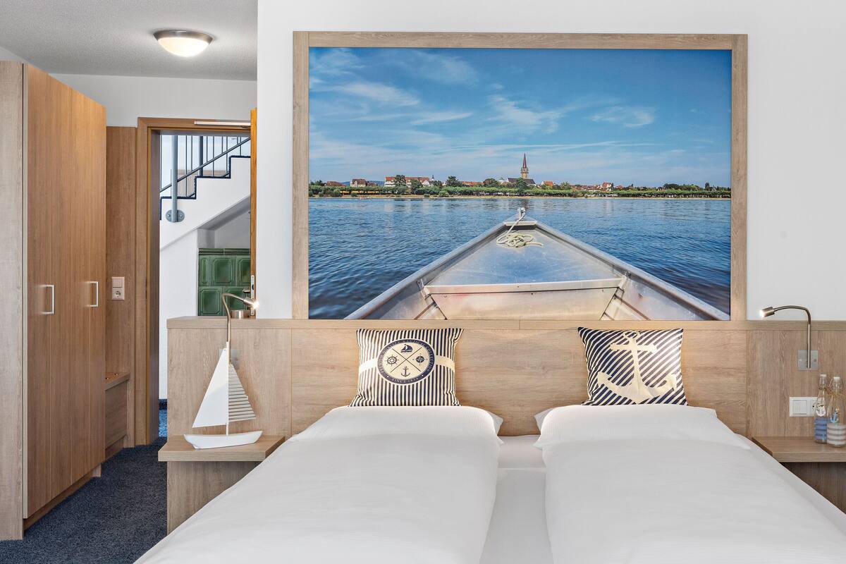 A hotel room with a bed, a wooden headboard, a painting of a boat on water, and a view of a building in the distance.