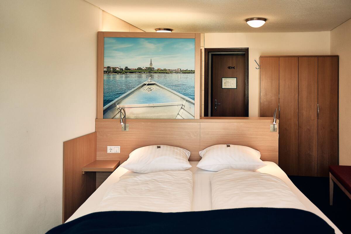 A hotel room with a bed, a wooden wardrobe, a door with a fish symbol, and a painting of a boat on the wall.