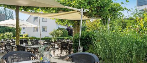 Un espace de restauration en plein air, avec des tables et des chaises disposées sous une tonnelle, entouré de verdure et avec un bâtiment en arrière-plan.