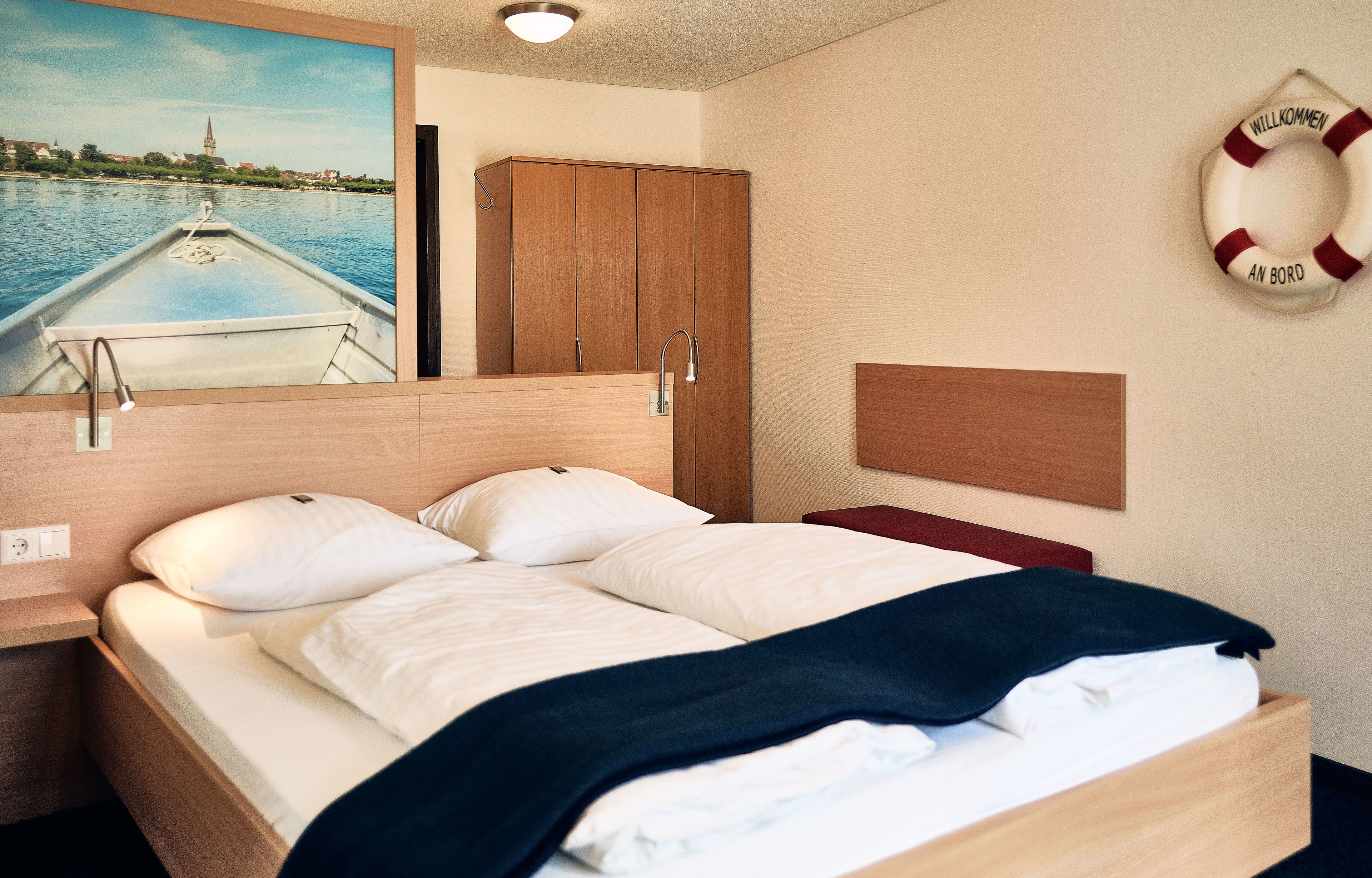 Une chambre d’hôtel avec un lit, une tête de lit en bois, un tableau représentant un paysage fluvial, un gilet de sauvetage accroché au mur et une armoire en bois.