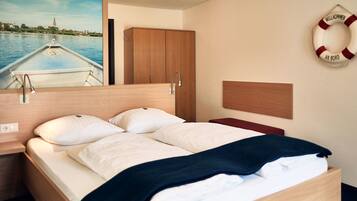Une chambre d’hôtel avec un lit, une tête de lit en bois, un tableau représentant un paysage fluvial, un gilet de sauvetage accroché au mur et une armoire en bois.