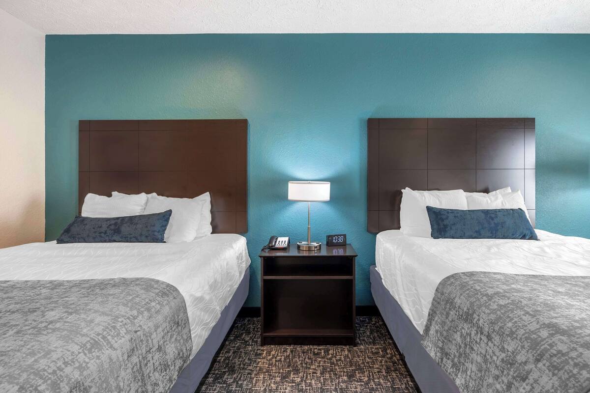 Habitación de hotel con dos camas, una mesita de noche con lámpara y un reloj que marca las 10:38.
