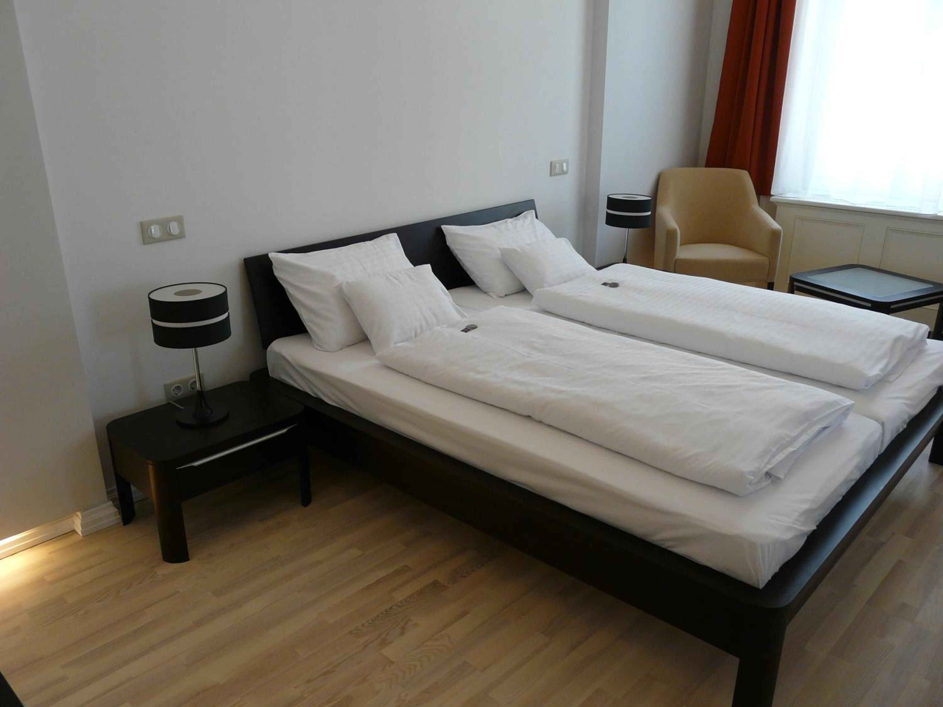 A double bed with white linens, a bedside table with a lamp, and a chair in a hotel room.