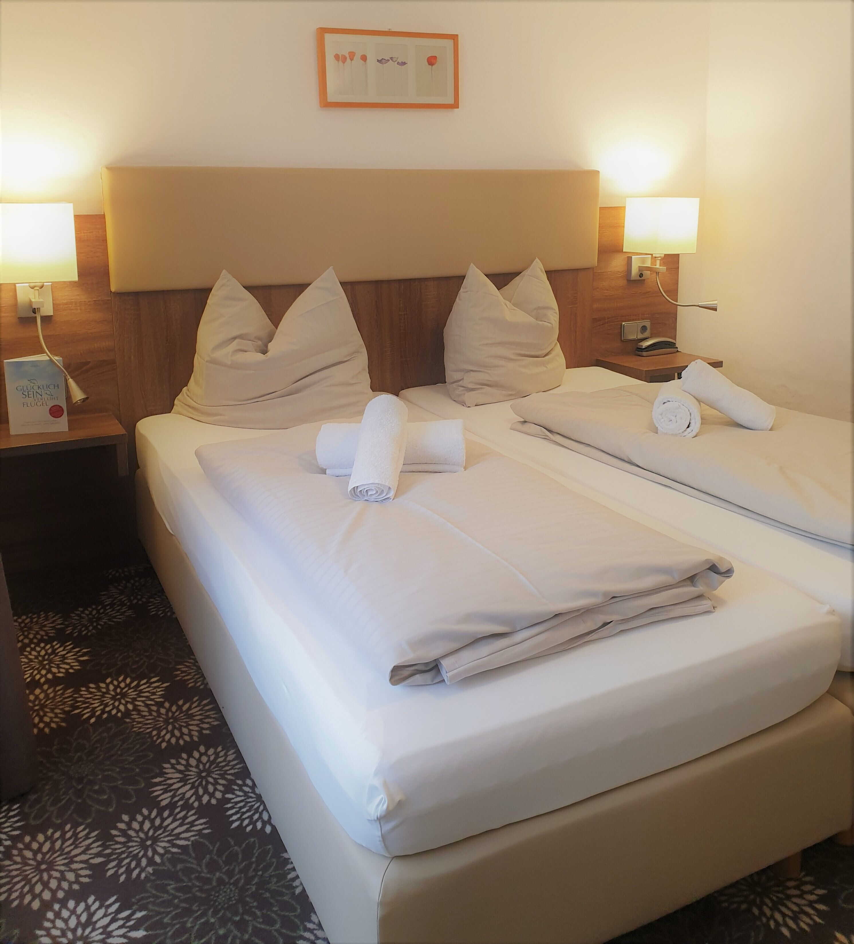 A hotel room with a double bed, two pillows, a bedside table with a lamp, and a framed picture on the wall.