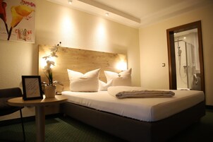 Quarto de hotel com cama, mesa de cabeceira, planta em vaso e área de chuveiro.