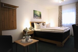 Quarto de hotel com uma cama, mesa de cabeceira, mesa com uma garrafa de água, quadro na parede e janela com cortinas.