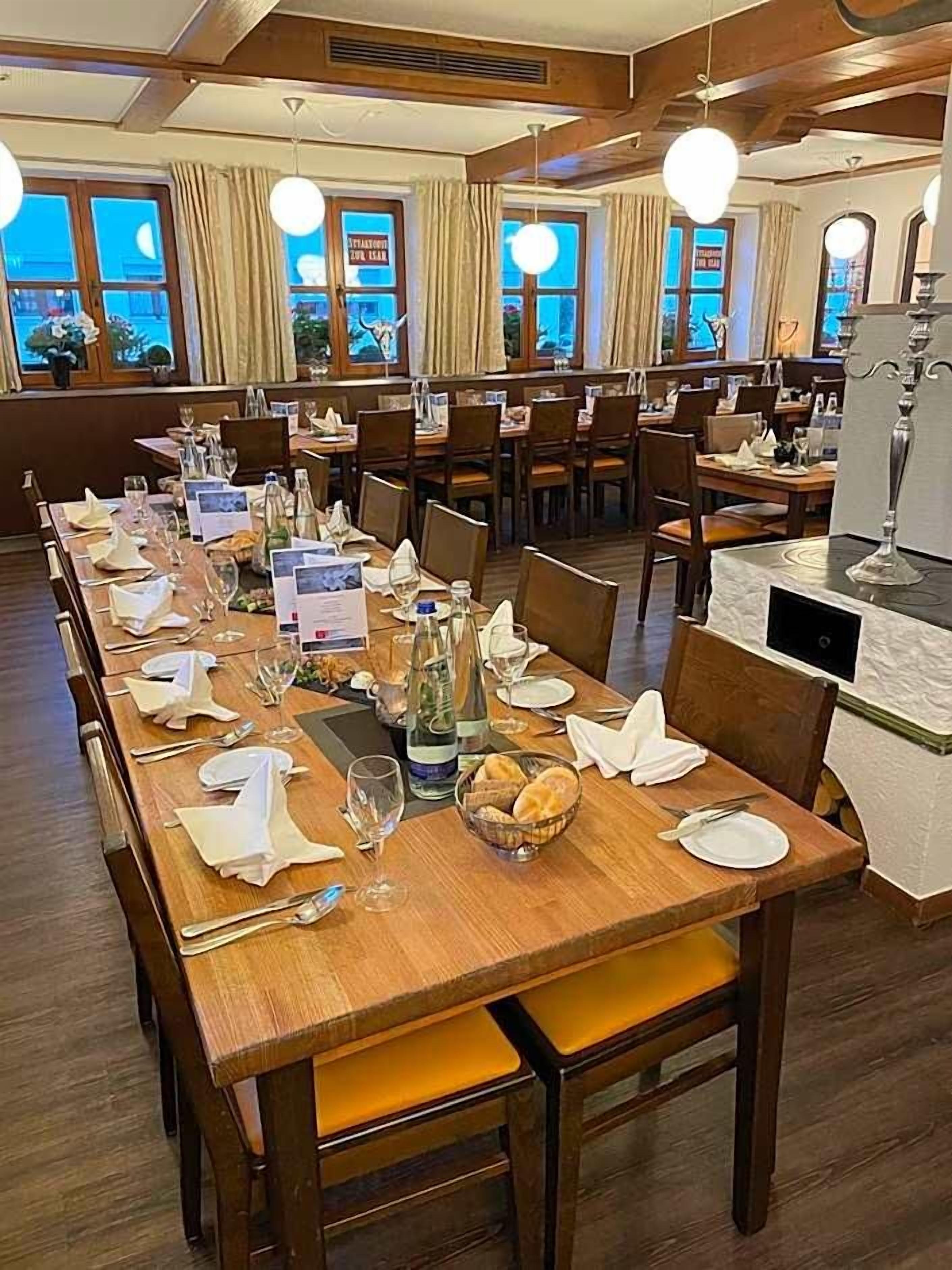 A restaurant with multiple dining tables set for service, wooden flooring, and large windows with curtains.