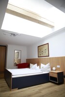 Quarto moderno com claraboia, cama com roupa de cama branca, cadeira vermelha, mesa de cabeceira com abajur e obra de arte emoldurada na parede.