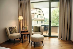 A room with two upholstered chairs, a small round table with a lamp, and a view of a house through a glass door. - Hofgut Farny (Kisslegg)