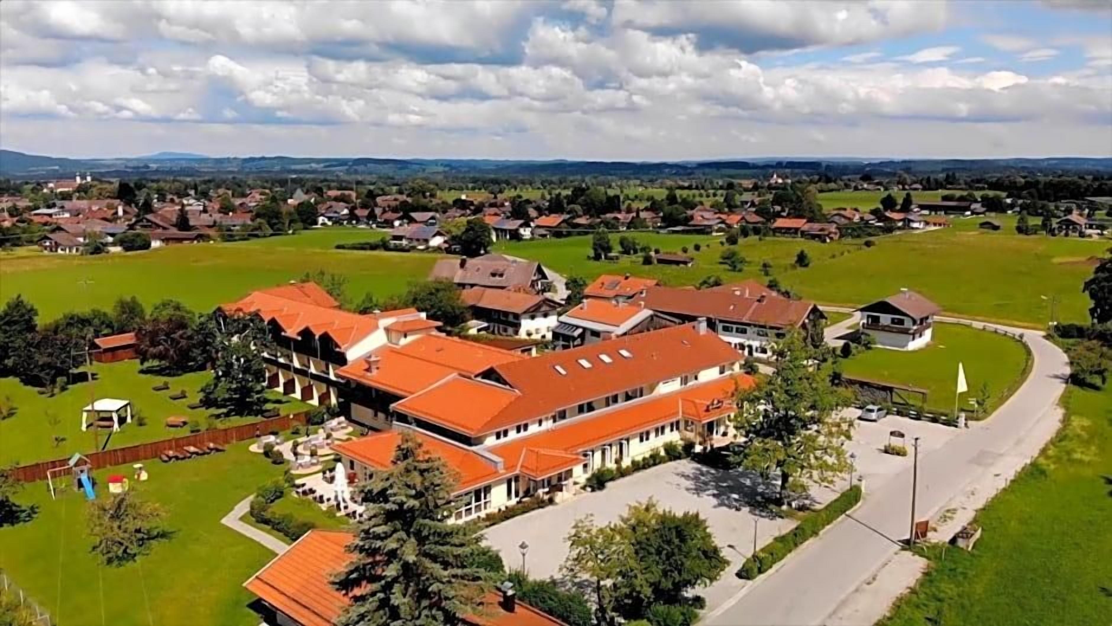 A large building with multiple red-roofed structures, surrounded by green fields and a few smaller buildings in the background.