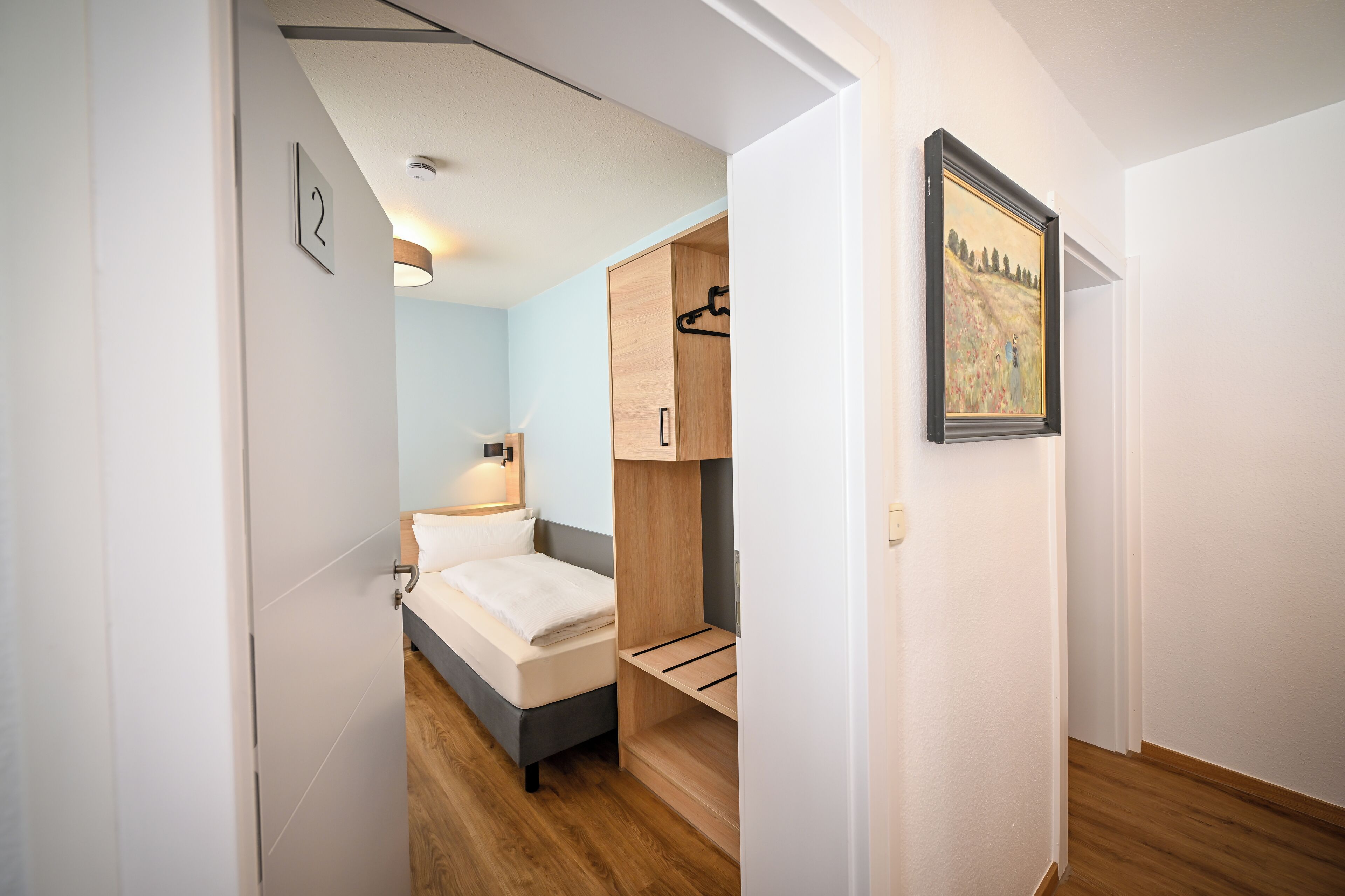 Un dormitorio con una cama, un armario de madera, un cuadro colgado en la pared y una puerta que da a otra habitación.