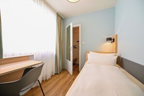 A hotel room with a wooden desk, a bed with white bedding, a wall-mounted lamp, and a view through an open door. - Hotel Mainblick (Marktheidenfeld)