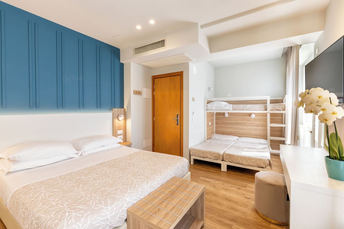 A modern hotel room with a large bed, a smaller bed, and a bunk bed. There is a wooden bench, a small table with a vase of flowers, and a TV on the wall.