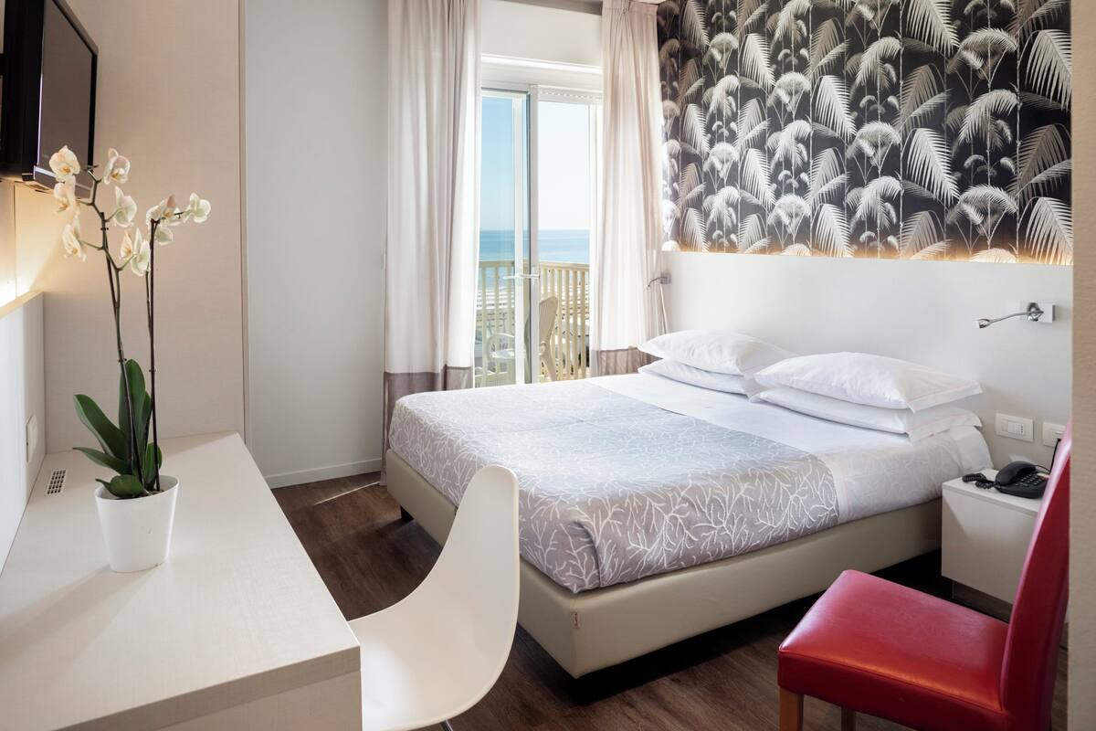 A hotel room with a bed, a desk, a chair, a potted plant, and a view of the ocean.