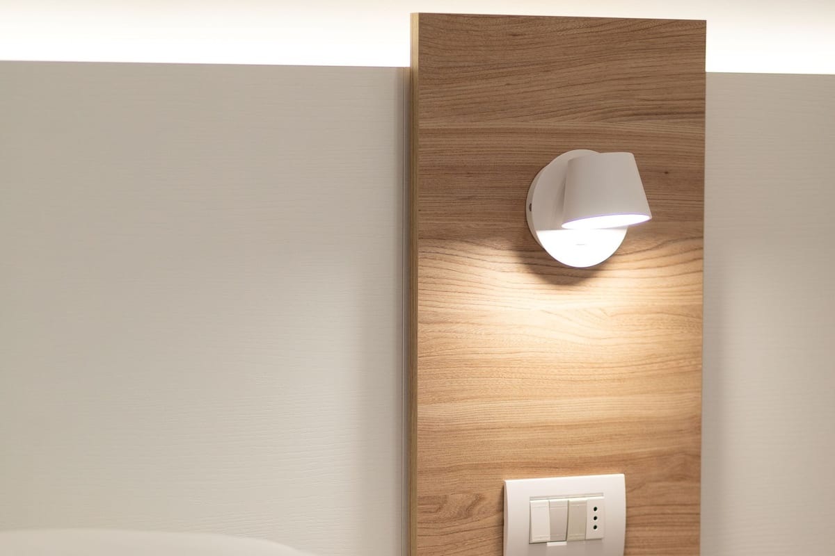 A neatly made bed with white linens, a wooden headboard, and a wall-mounted light fixture.