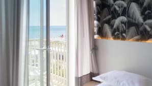 Quarto de hotel com varanda e vista para a praia, cama com lençóis brancos e parede com padrão de folha de palmeira.