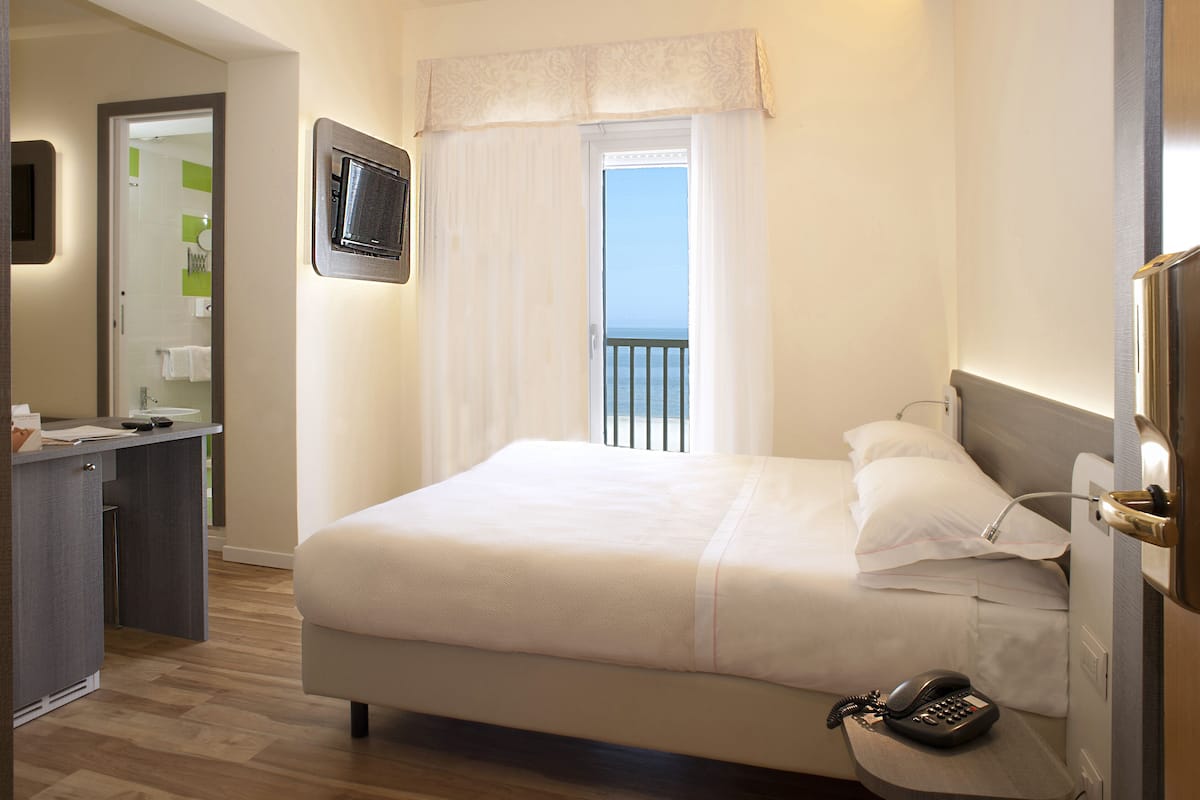 A hotel room with a bed, a desk, a telephone, and a window with a view of the sky.