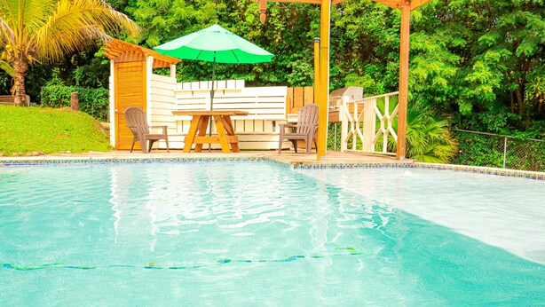 A swimming pool with a wooden deck, two chairs, and a green umbrella. - Forst Villa (Kingstown)