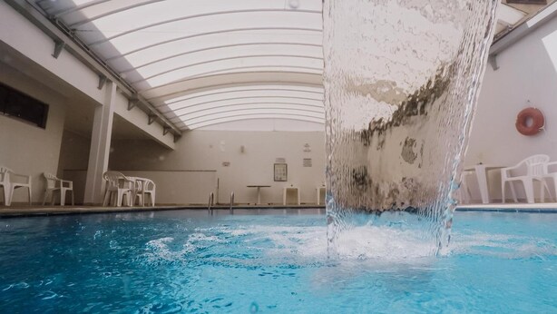 A swimming pool with a water fountain in the center, surrounded by white chairs and a curved ceiling structure. - Alzira Granada Hotel (Sao Lourenco)