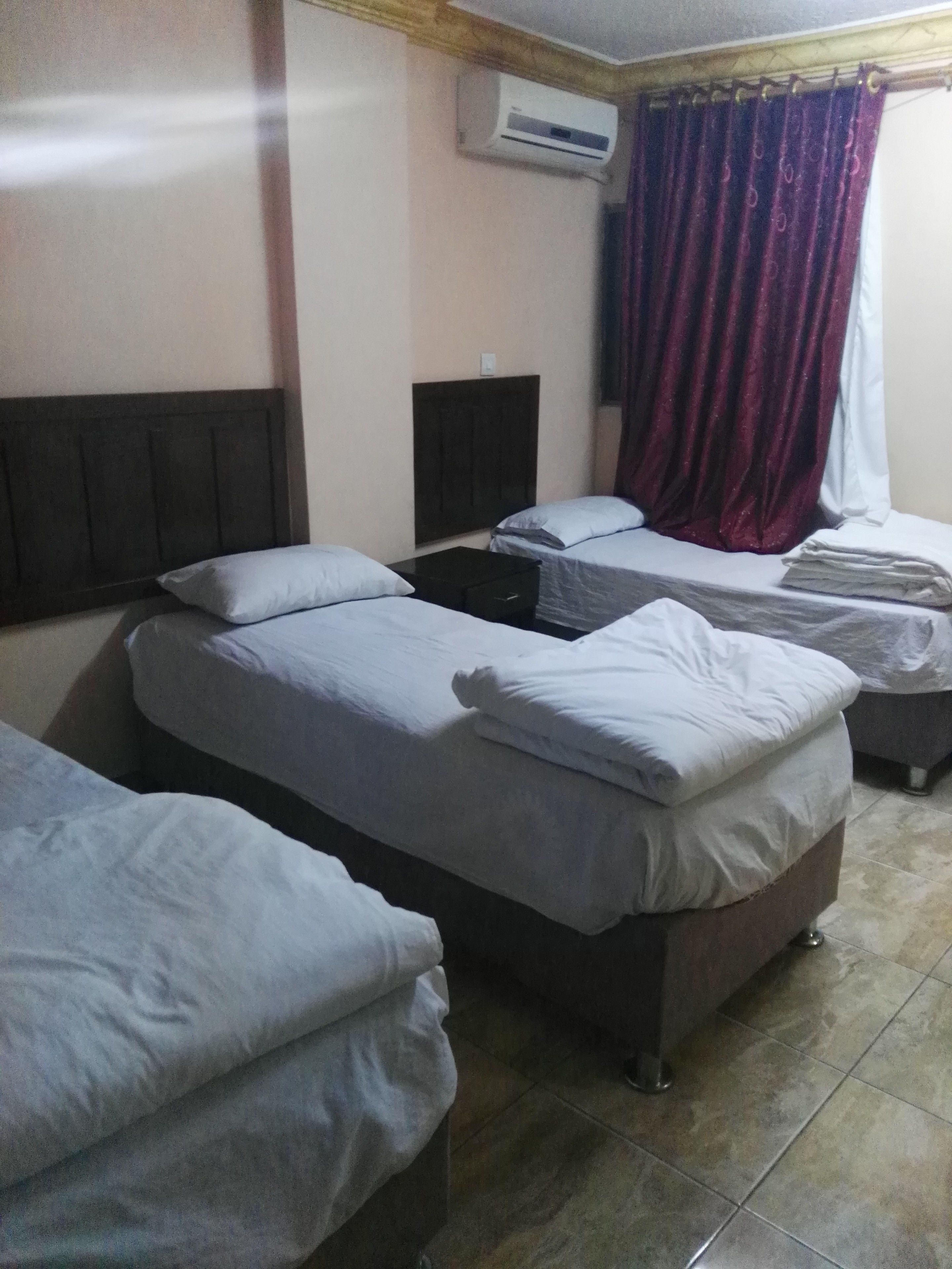 Una camera d'albergo con tre letti singoli, una testiera scura, un condizionatore d'aria e una finestra con tenda.