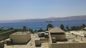 Vista costeira com um grande navio ao longe, céu claro e um edifício com antenas parabólicas.