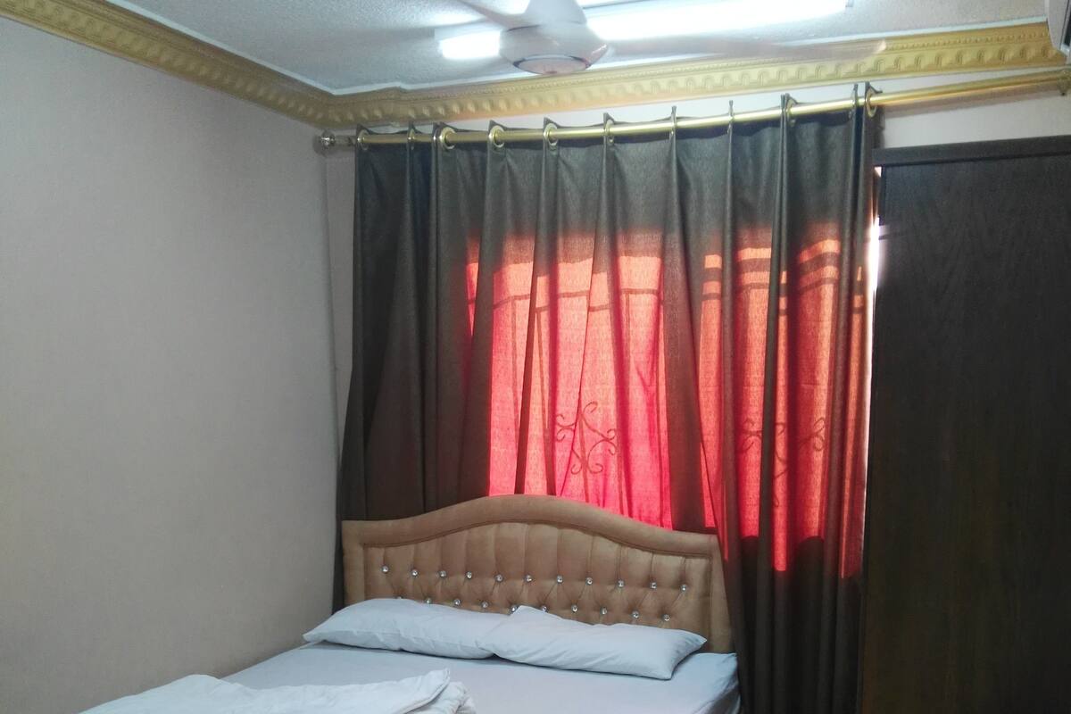 A neatly made bed with a headboard, a ceiling fan, and a window with red curtains.