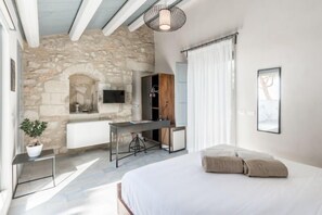 Une chambre avec un mur en pierre, un lit, un bureau, une télévision et une fenêtre avec des rideaux.