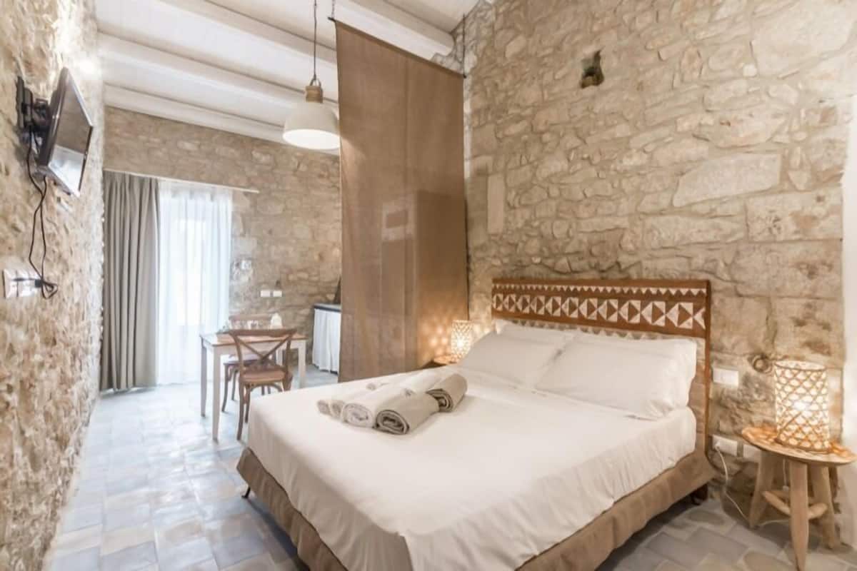 A bedroom with a stone wall, a bed with rolled towels, a wooden headboard, a small table with a lamp, and a TV mounted on the wall.