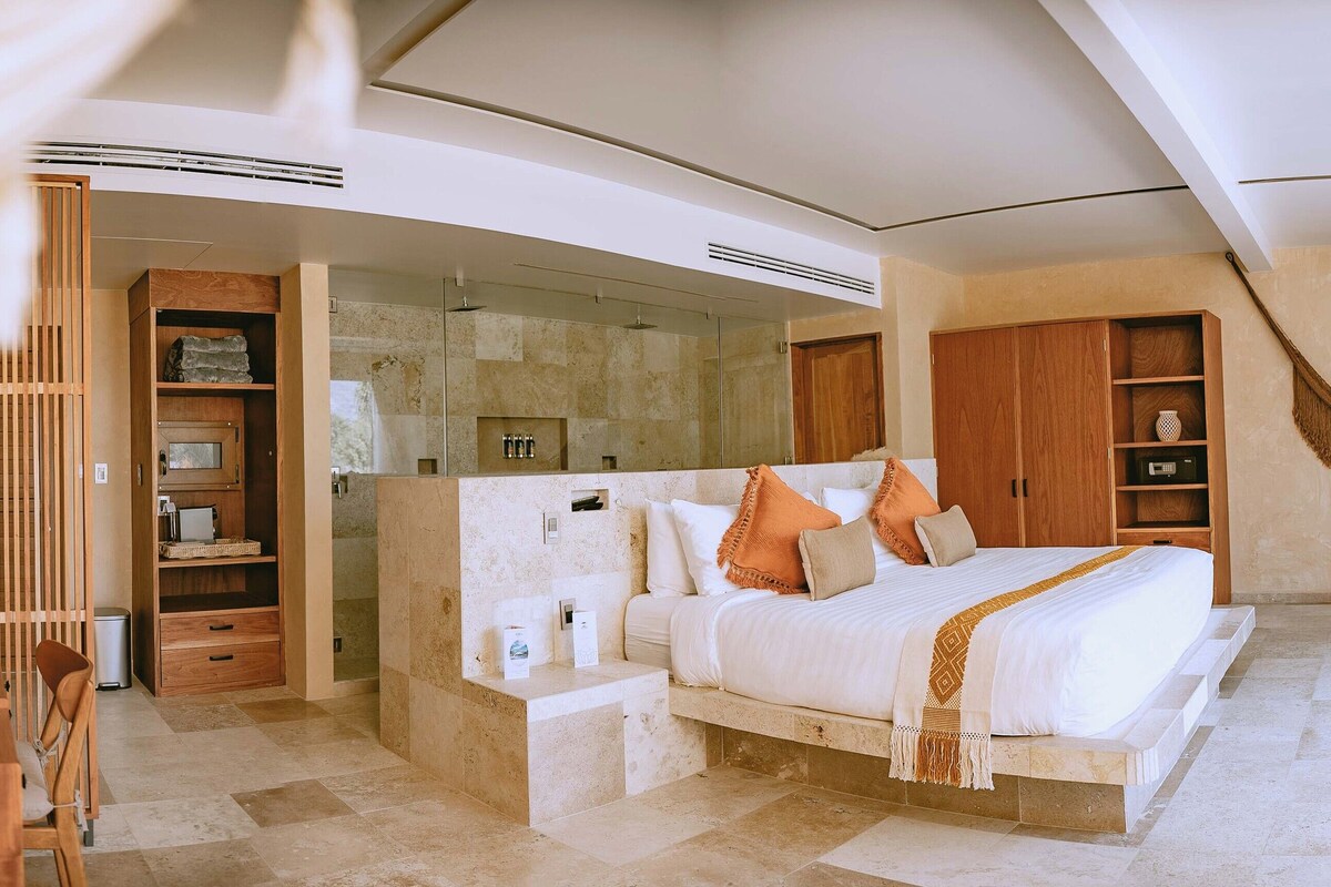 A modern bedroom with a large bed, wooden furniture, a glass shower, and a hanging macrame piece.