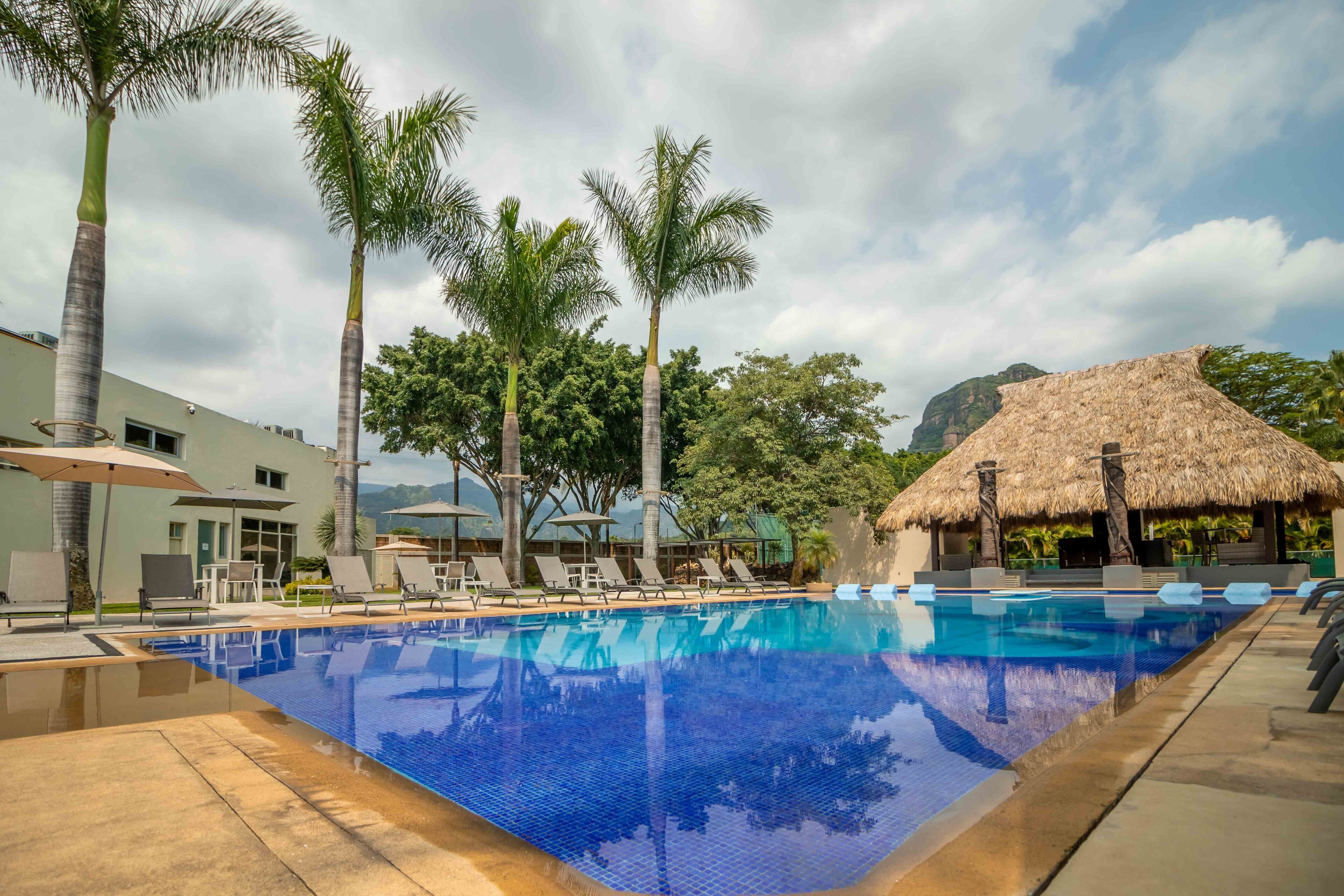 Una piscina con sillas de descanso y sombrillas, rodeada de palmeras y una estructura con techo de paja.