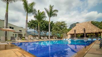 Una piscina con sillas de descanso y sombrillas, rodeada de palmeras y una estructura con techo de paja.