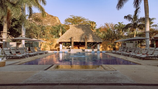 A pool area with lounge chairs, a thatched-roof structure, and palm trees.