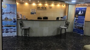 A reception area with a marble floor, two chairs, a reception desk, and informational posters. - Ahla Tlah SeaView Hotel (Aqaba)