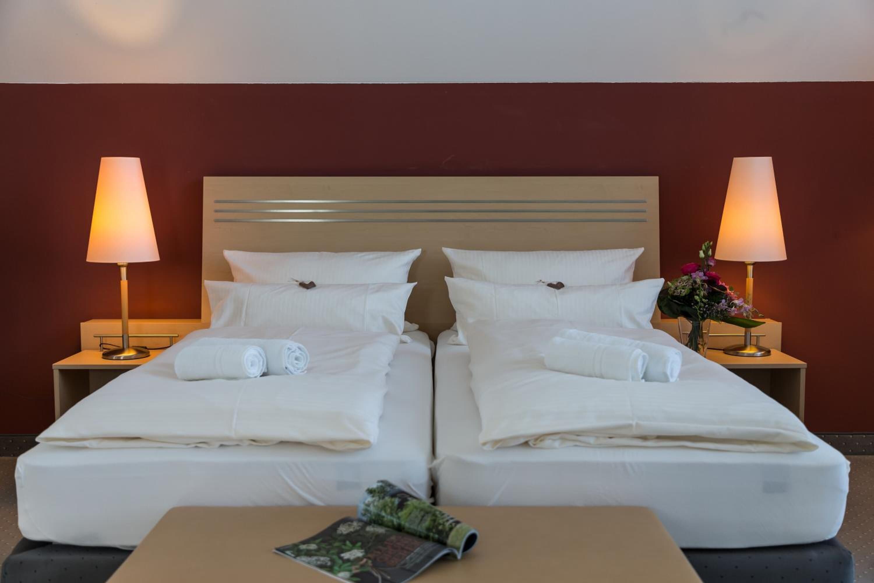 Habitación de hotel con dos camas, cada una con sábanas y almohadas blancas, cabeceros de madera, lámparas de noche y una mesita con una revista.