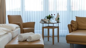 Et hotelværelse med en seng, to stole, et lille bord med en vase og en flaske, samt et vindue med let gardin.