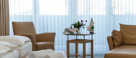 Une chambre d’hôtel avec un lit, deux chaises, une petite table avec un vase et une bouteille, et une fenêtre avec des rideaux légers.