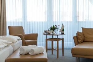 Quarto de hotel com cama, duas cadeiras, uma mesinha com um vaso e uma garrafa, e uma janela com cortinas finas.