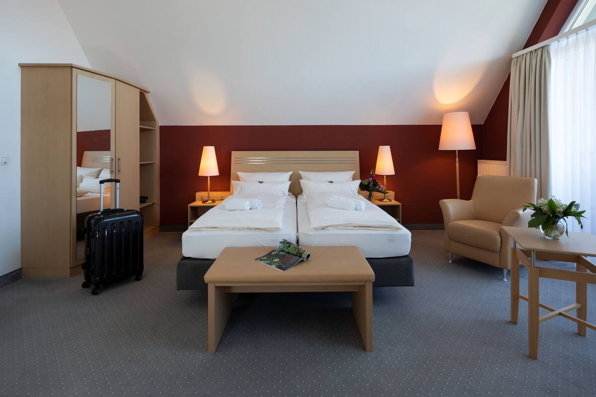 A hotel room with a bed, two bedside tables with lamps, a chair, a small table, a desk, a mirror, and a suitcase.