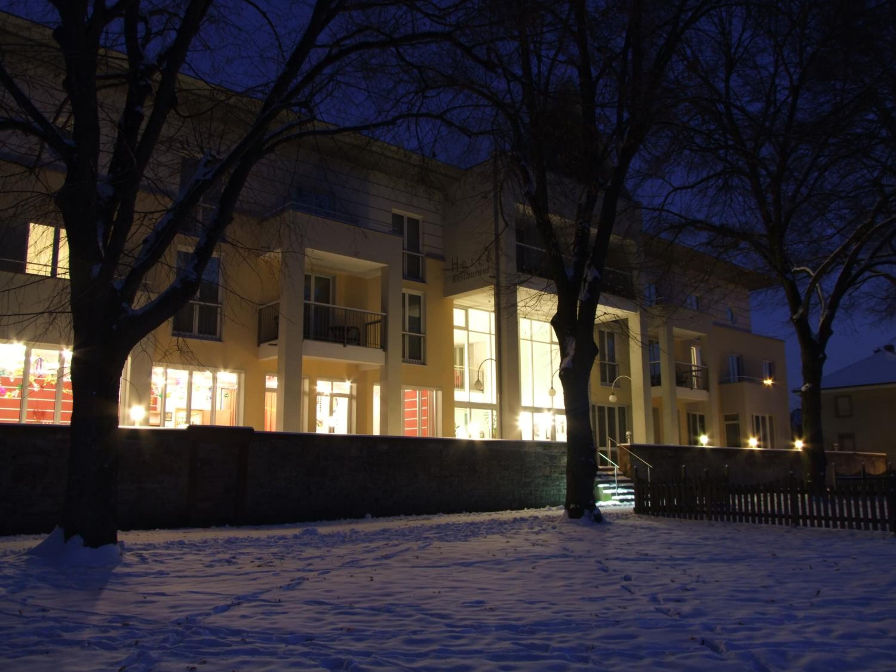 Monikerroksinen rakennus, jonka ikkunat on valaistu ja jonka edusta on lumen peitossa.