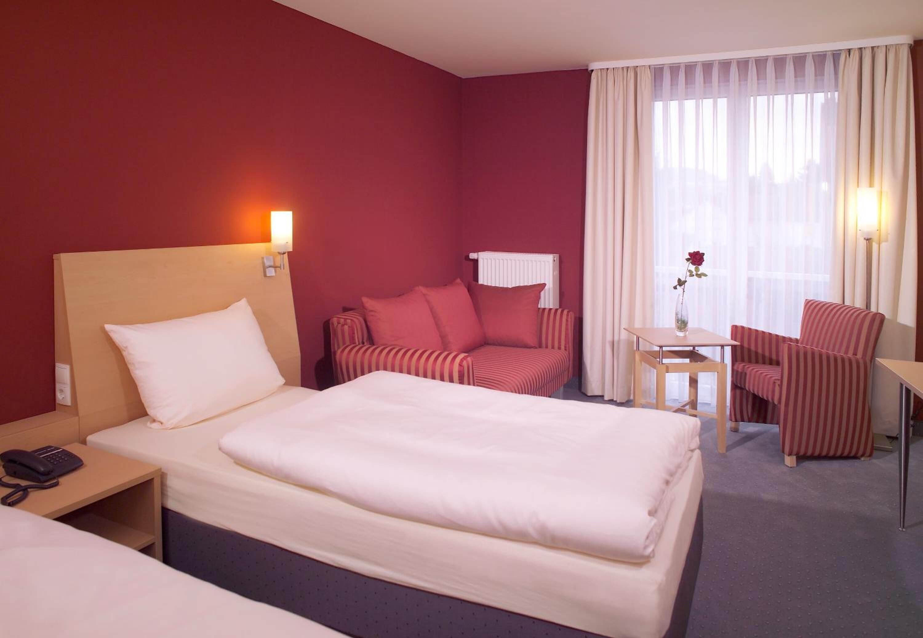 A hotel room with a large bed, a smaller bed, a sofa, a chair, a table, and a vase with a flower.